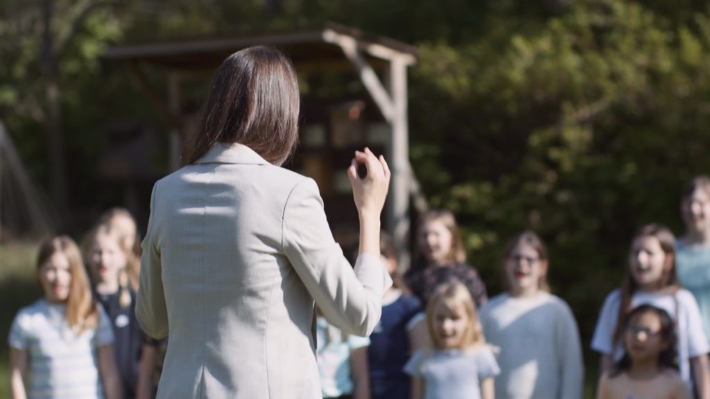Zu sehen ist eine langhaarige Person von hinten, die einen Kinderchor unter freiem Himmel dirigiert.