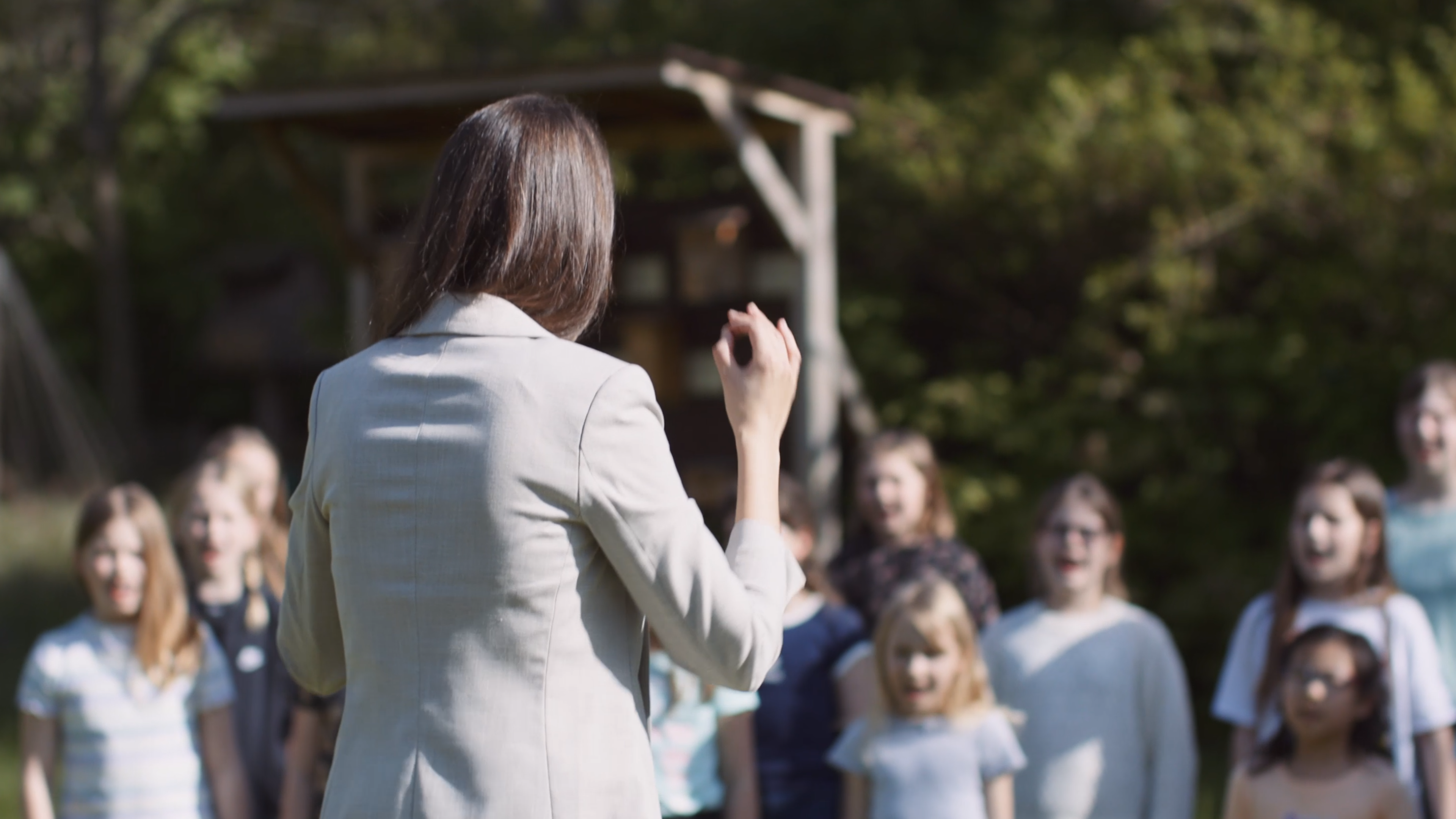 Zu sehen ist eine langhaarige Person von hinten, die einen Kinderchor unter freiem Himmel dirigiert.